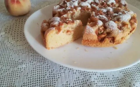 Delizioso ciasto z brzoskwiniami, cosparso di zucchero a velo, su un piatto bianco, accanto a una pesca fresca.