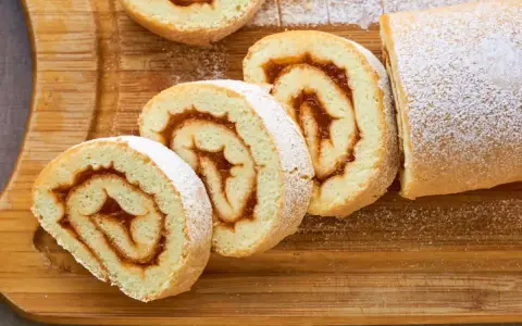Rotoli di pasta biscotto 3 uova con ripieno di marmellata, spolverati di zucchero a velo su tagliere di legno.