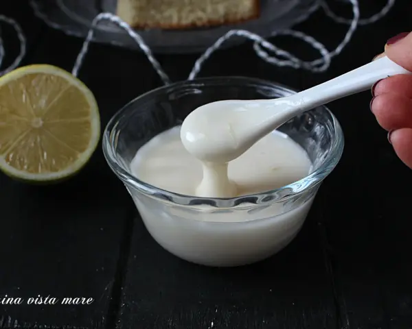 Una mano preleva con un cucchiaio una densa glassa al limone trasparente da una ciotola di vetro. Accanto, una fetta di limone.