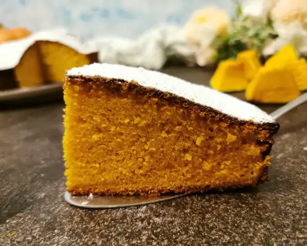 Una fetta di torta zucca senza glutine, soffice e dorata, cosparsa di zucchero a velo. Sullo sfondo, pezzi di zucca e un'altra torta.