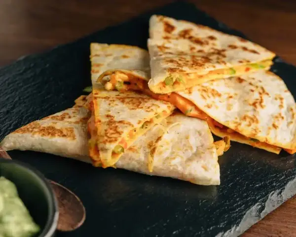 Deliziose quesadillas di pollo dorate e ripiene di formaggio filante, servite su una pietra scura con una ciotola di guacamole.
