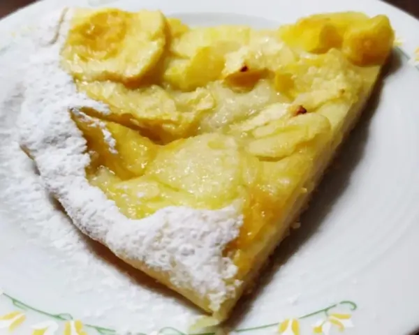Una fetta di pizza con le mele, cosparsa di zucchero a velo, su un piatto bianco con decorazioni floreali.
