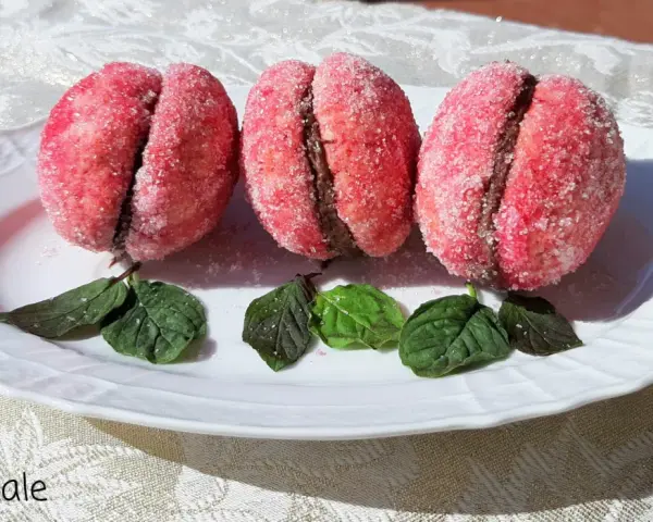 Tre dolci pesche abruzzesi ricoperte di zucchero, con un ripieno scuro, adagiate su un piatto bianco con foglie verdi.