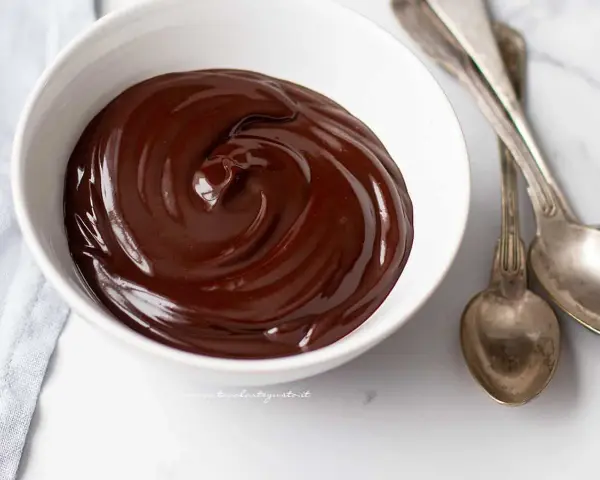 Una ciotola bianca piena di glassa al cioccolato e burro, lucida e invitante, con due cucchiai d'argento accanto.