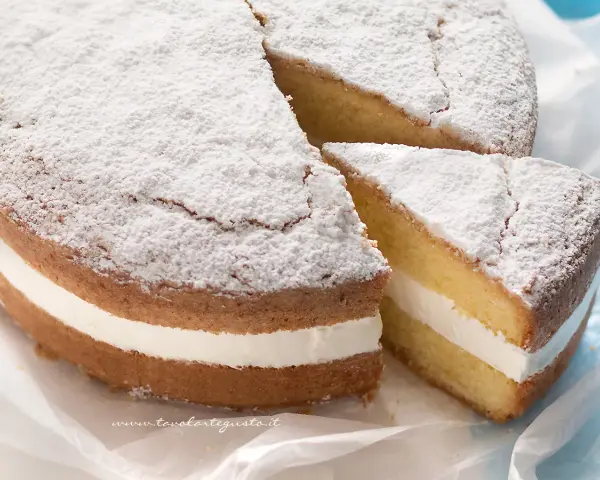 Una soffice torta paradiso con crema bianca, spolverata di zucchero a velo. Una fetta è stata tagliata, rivelando la deliziosa farcitura.