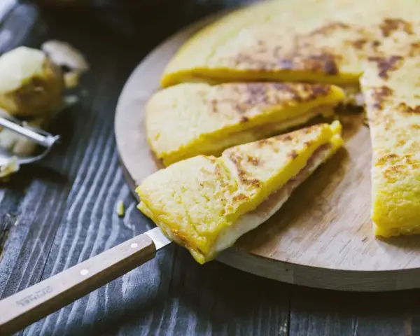 Focaccia di patate in padella con prosciutto cotto e formaggio, tagliata a spicchi su un tagliere di legno.