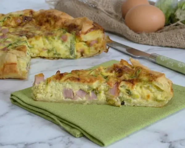 A slice of ham and zucchini quiche on a green napkin, with the rest of the quiche in the background.