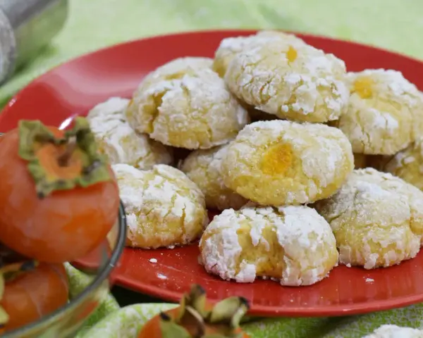 Biscotti ai cachi, dolcetti morbidi e profumati, cosparsi di zucchero a velo, accanto a cachi freschi.