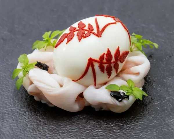 Uovo sodo decorato con motivi rossi, adagiato su un letto di calamari crudi e guarnito con erbe aromatiche. Un barzotto uovo elegante.