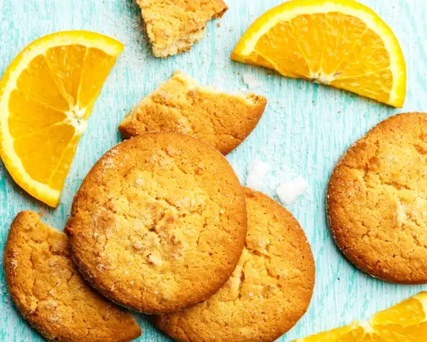 Biscotti all'arancia ricetta della nonna: dolci profumati e dorati, con spicchi d'arancia fresca su un tavolo di legno azzurro.