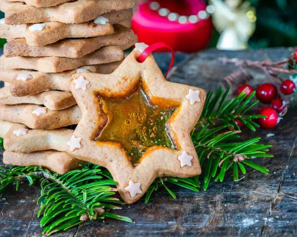 Biscotti a forma di stella con centro trasparente, decorati con piccoli fiocchi di neve, circondati da rami di abete e bacche rosse.
