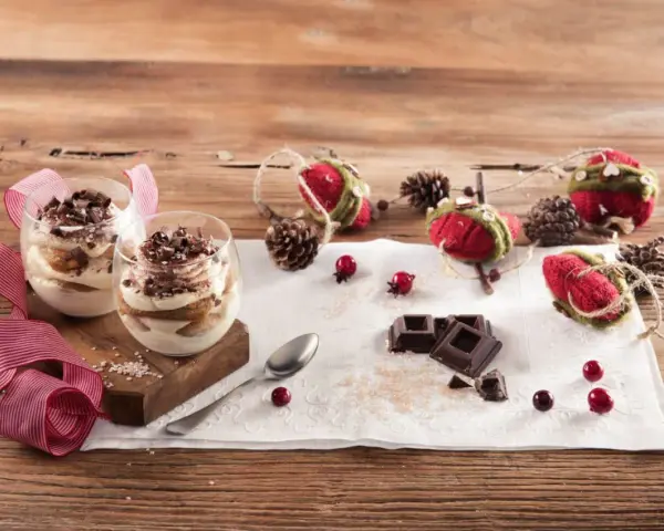 Due bicchieri di tiramisù Galbani, decorati con cioccolato e nastri rossi, su un tavolo di legno con decorazioni natalizie.