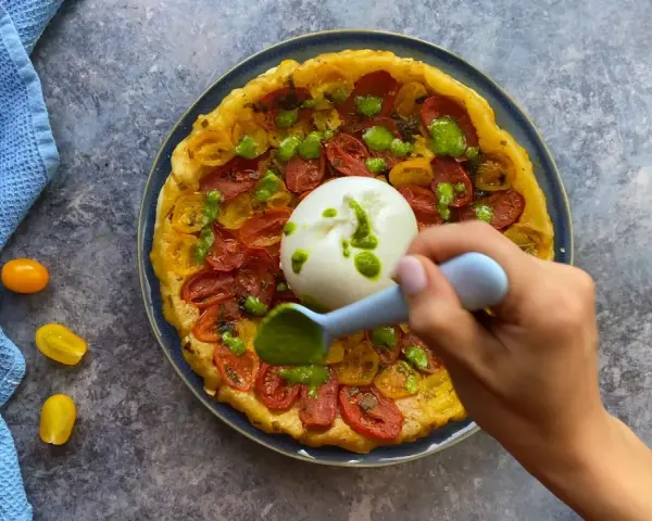 Tarte tatin pomodorini e burrata, con una mano che aggiunge un pesto verde.