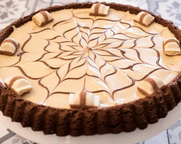 Torta con base croccante al cacao, ripiena di crema chiara e decorata con cioccolato fondente e pezzi di Kinder Bueno White.