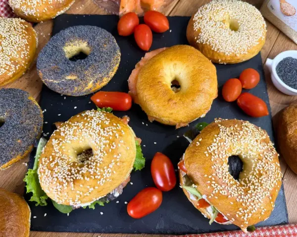 Varietà di bagel dolci e salati, alcuni con semi di sesamo, altri con semi di papavero, accompagnati da pomodorini e salmone affumicato.