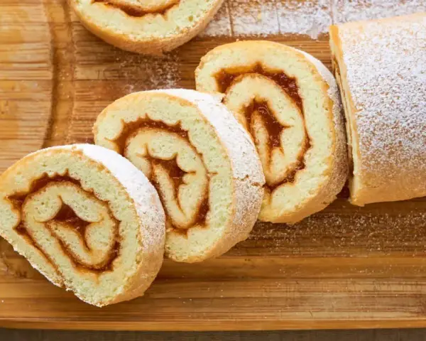 Rotoli di pasta biscotto 3 uova con ripieno di marmellata, spolverati di zucchero a velo su tagliere di legno.