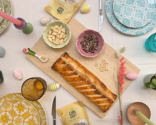 Torta pasqualina senza uova dorata, con mandorle e pinoli, decorata con uova colorate e candela pasquale.