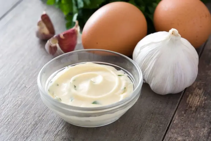 Una ciotola di cremoso sos aioli, con uova, aglio e prezzemolo sullo sfondo.