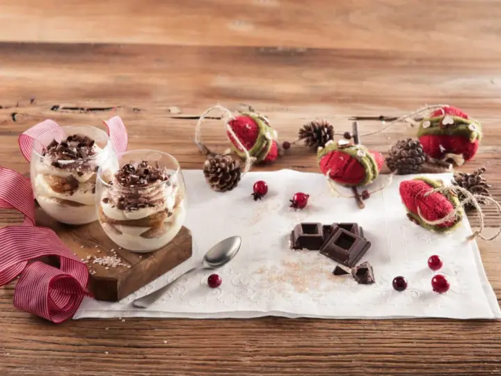Due bicchieri di tiramisù Galbani, decorati con cioccolato e nastri rossi, su un tavolo di legno con decorazioni natalizie.