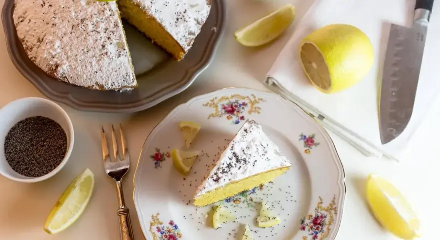 Una fetta di babka cytrynowa, cosparsa di zucchero a velo e semi di papavero, con spicchi di limone e una ciotolina di semi.