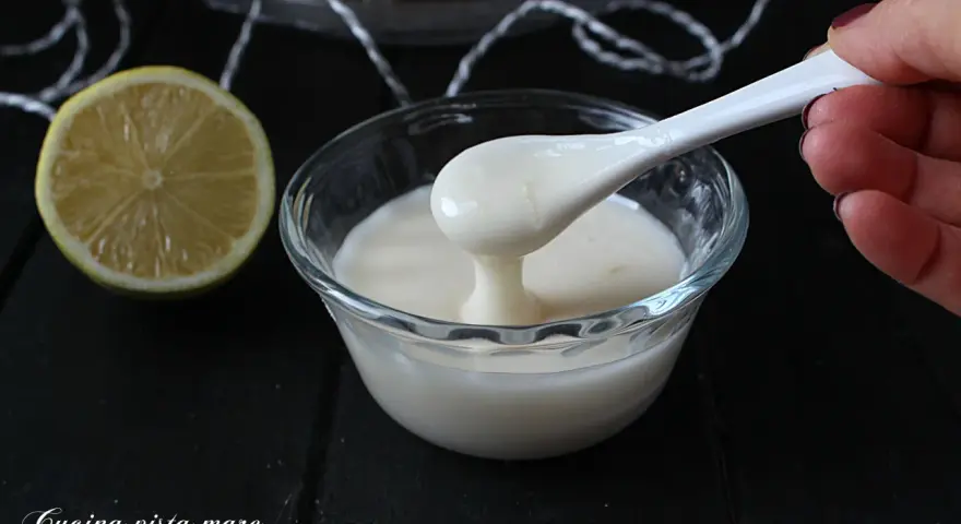 Una mano preleva con un cucchiaio una densa glassa al limone trasparente da una ciotola di vetro. Accanto, una fetta di limone.