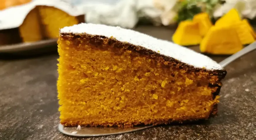 Una fetta di torta zucca senza glutine, soffice e dorata, cosparsa di zucchero a velo. Sullo sfondo, pezzi di zucca e un'altra torta.