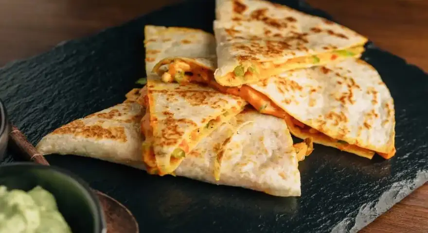 Deliziose quesadillas di pollo dorate e ripiene di formaggio filante, servite su una pietra scura con una ciotola di guacamole.
