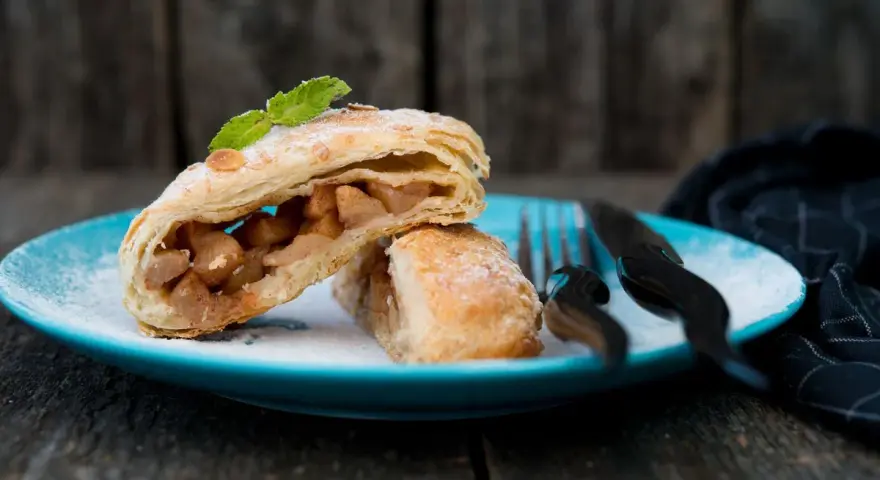 Delizioso strudel pere cioccolato e amaretti, con ripieno di mele e un tocco di menta, su un piatto blu cosparso di zucchero a velo.