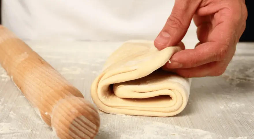 Mano che piega un impasto stratificato, pronto per diventare delizioso **ciasto francuskie**. Un mattarello è accanto.