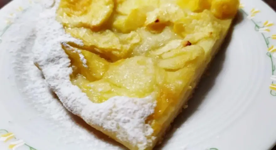 Una fetta di pizza con le mele, cosparsa di zucchero a velo, su un piatto bianco con decorazioni floreali.