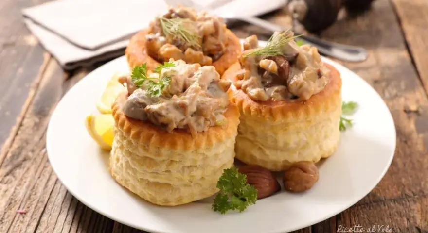 Tre vol-au-vent con funghi cremosi, guarniti con prezzemolo e aneto, su un piatto bianco con spicchi di limone.
