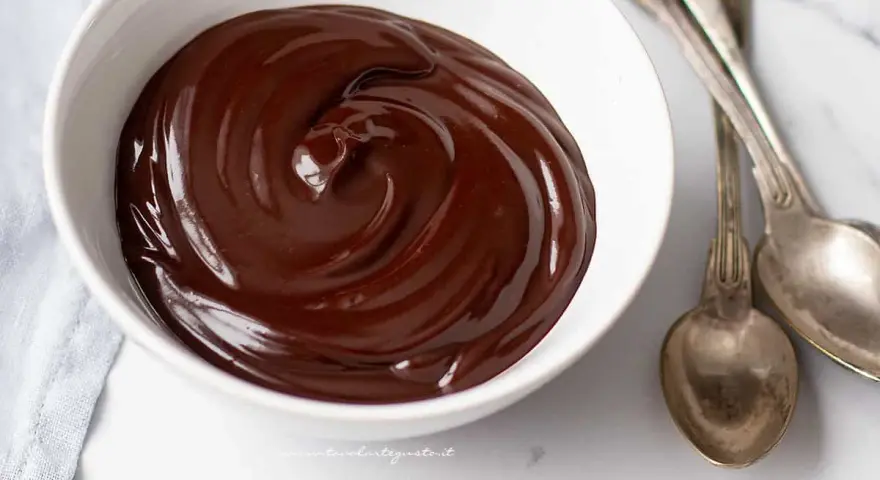 Una ciotola bianca piena di glassa al cioccolato e burro, lucida e invitante, con due cucchiai d'argento accanto.