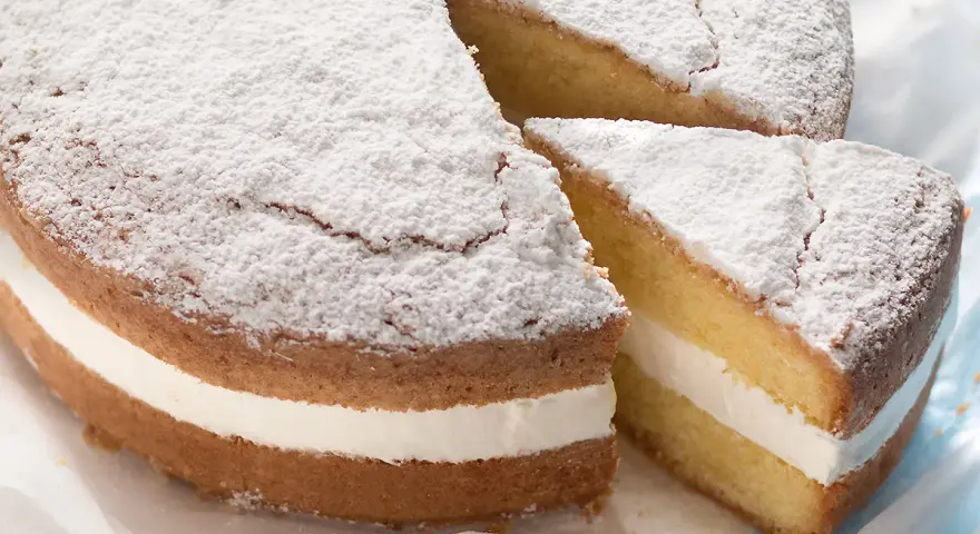 Una soffice torta paradiso con crema bianca, spolverata di zucchero a velo. Una fetta è stata tagliata, rivelando la deliziosa farcitura.