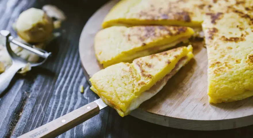 Focaccia di patate in padella con prosciutto cotto e formaggio, tagliata a spicchi su un tagliere di legno.