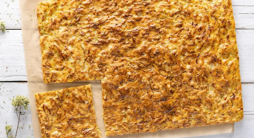 Schiacciata di zucchine e cipolle dorata e croccante, tagliata a pezzi su carta da forno.