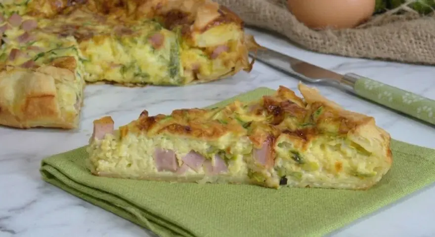 A slice of ham and zucchini quiche on a green napkin, with the rest of the quiche in the background.