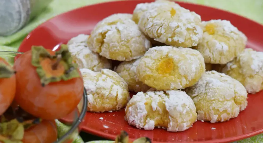 Biscotti ai cachi, dolcetti morbidi e profumati, cosparsi di zucchero a velo, accanto a cachi freschi.