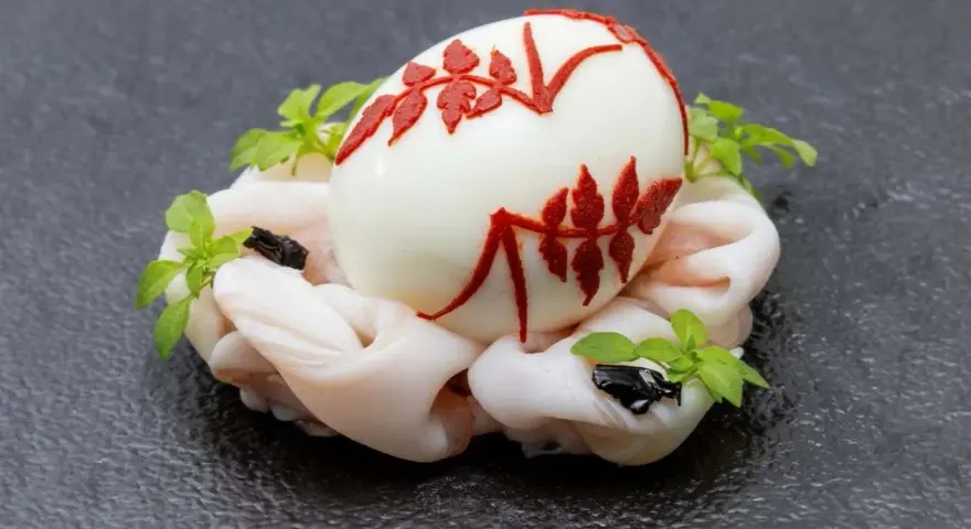 Uovo sodo decorato con motivi rossi, adagiato su un letto di calamari crudi e guarnito con erbe aromatiche. Un barzotto uovo elegante.