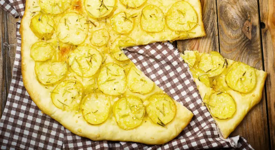 Schiacciata di patate con farina di ceci, guarnita con rosmarino fresco, su un tagliere di legno rustico.