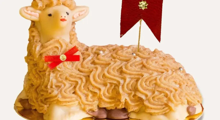 A decorative cake shaped like a lamb, with piped frosting and a red ribbon.