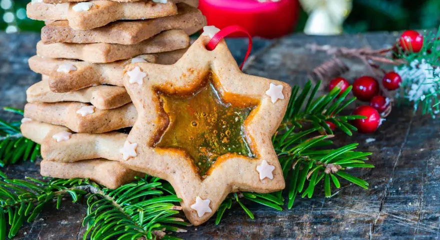 Biscotti a forma di stella con centro trasparente, decorati con piccoli fiocchi di neve, circondati da rami di abete e bacche rosse.