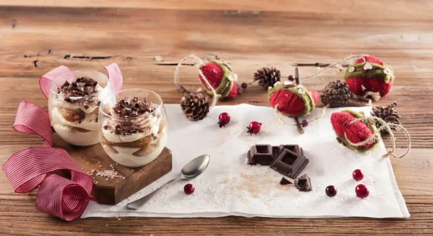 Due bicchieri di tiramisù Galbani, decorati con cioccolato e nastri rossi, su un tavolo di legno con decorazioni natalizie.