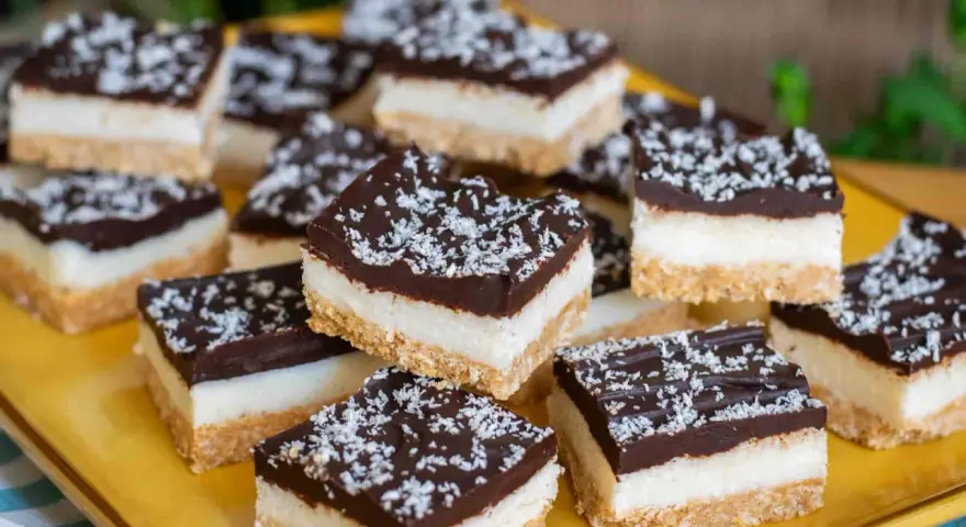 Deliziosi quadrotti cioccolato e cocco, stratificati con base croccante, crema bianca e copertura fondente, spolverati con cocco grattugiato.