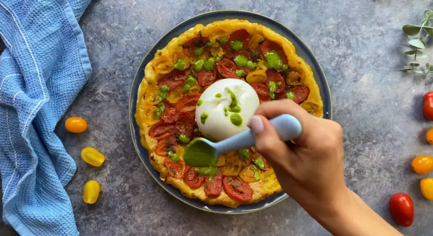 Tarte tatin pomodorini e burrata, con una mano che aggiunge un pesto verde.