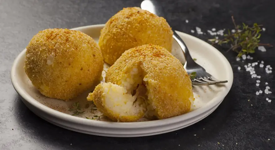Tre arancini dorati su un piatto, uno aperto rivela il ripieno di riso. Un invito a provare il nostro arancini ricetta.