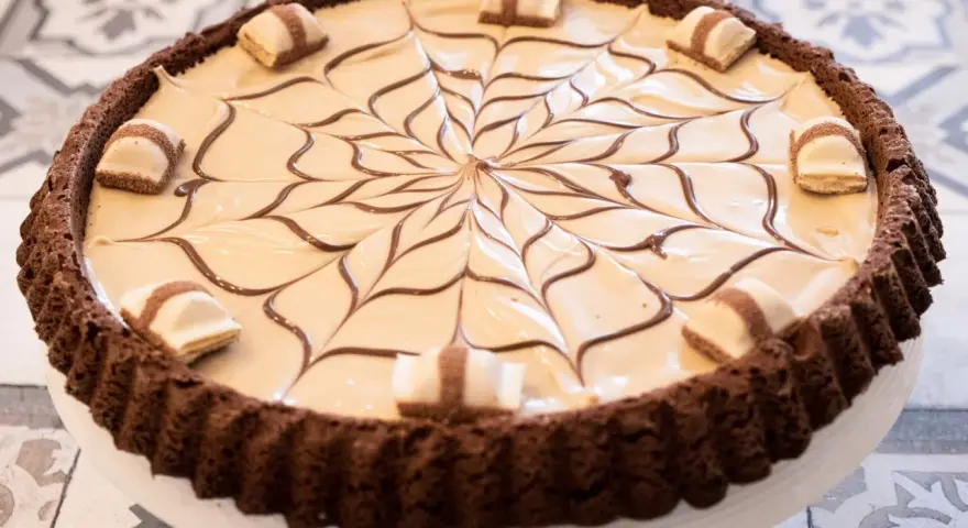 Torta con base croccante al cacao, ripiena di crema chiara e decorata con cioccolato fondente e pezzi di Kinder Bueno White.