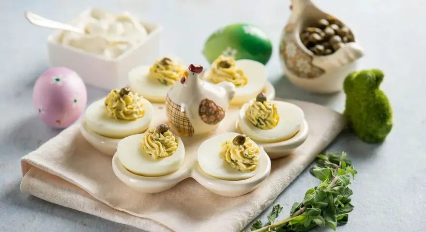 Uova ripiene vegetariane con ceci, guarnite con capperi e erbe aromatiche, su un piatto decorato con galline e uova pasquali.