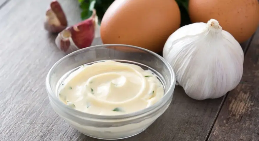 Una ciotola di cremoso sos aioli, con uova, aglio e prezzemolo sullo sfondo.