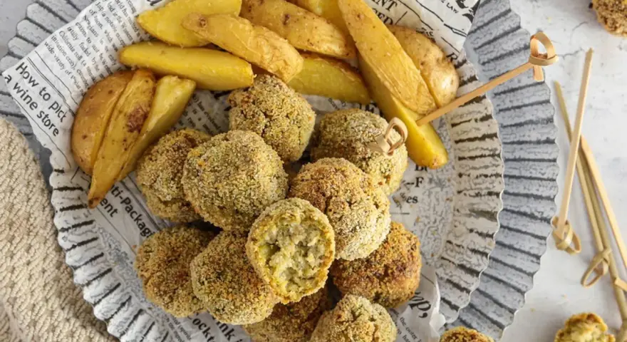 Polpette di verza e ricotta dorate e croccanti, servite con spicchi di patate fritte su carta di giornale.