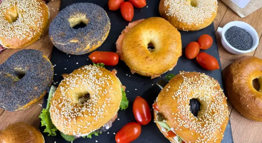 Varietà di bagel dolci e salati, alcuni con semi di sesamo, altri con semi di papavero, accompagnati da pomodorini e salmone affumicato.